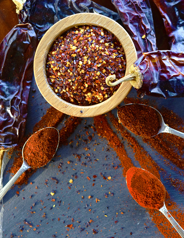 An overhead shot of red pepper flakes in a wooden bowl surrounded by chili powders on spoons