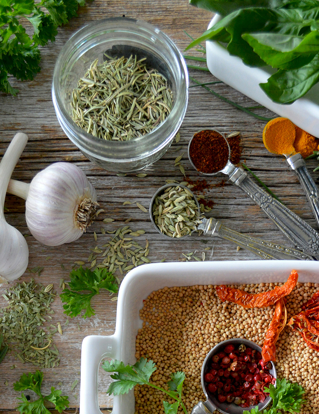 A flat-lay arrangement of various whole spices, fresh garlic, and green herbs on a wooden table