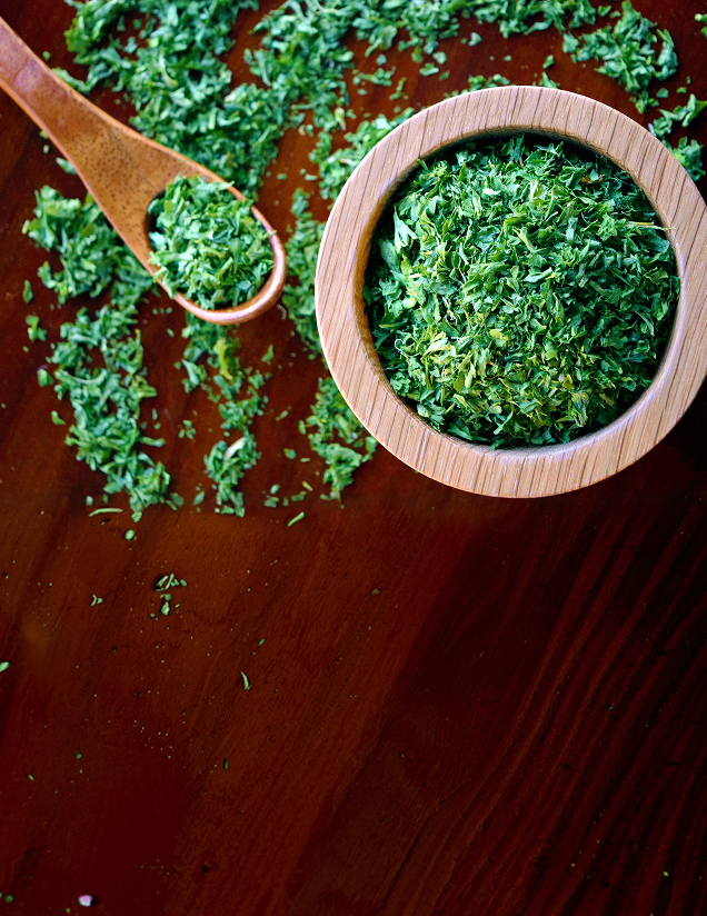 A wooden bowl and spoon filled with vibrant green dried parsley on a dark wood surface
